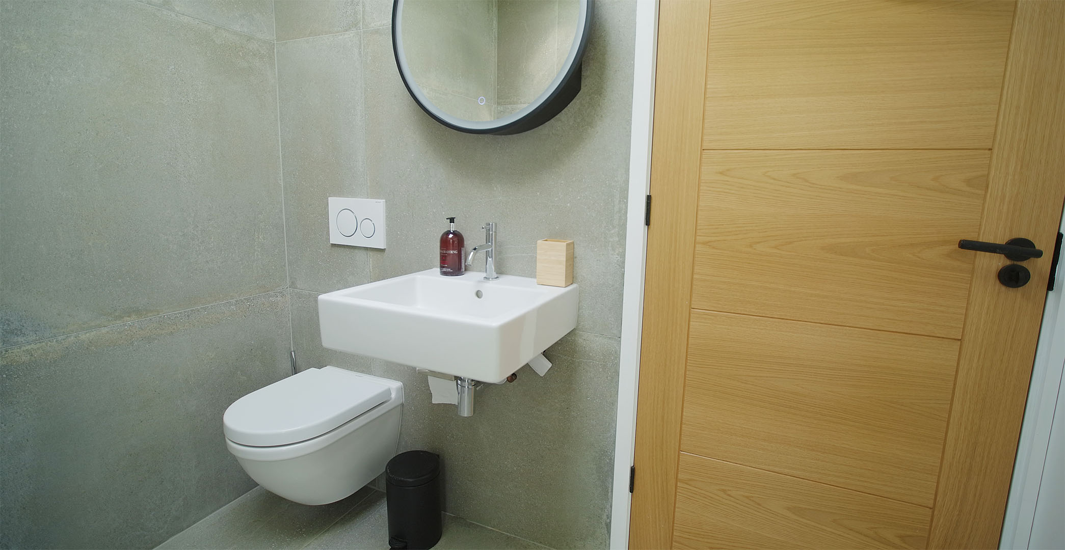 The wet room at Fernleigh Courtyard luxury holiday home showing grey tiles, wash basin, illuminated mirror cabinet, & toilet.